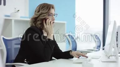 年轻的休闲<strong>女</strong>士坐在<strong>办公</strong>桌前，用笔记本电脑，<strong>接电话</strong>，打<strong>电话</strong>，微笑着在手机上说话